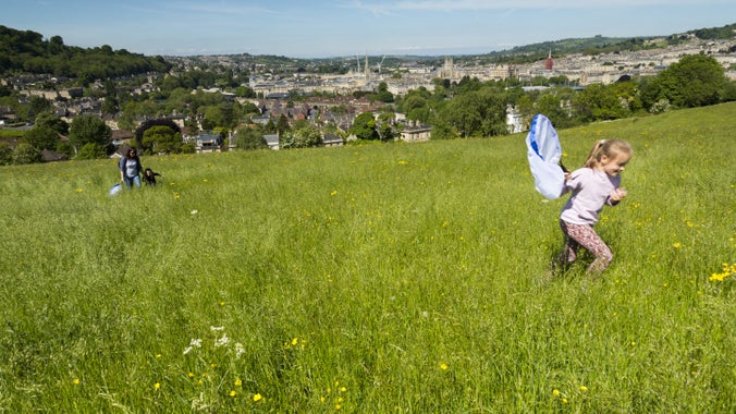 A family in a wildflower meadow running and holding nets to catch bugs. City of Bath in the distance.